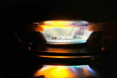 A close-up view of a gas stove burner with a bright blue flame, accented by hints of orange and yellow. The metal components of the burner are visible around the flame, and the surrounding area is dimly lit.