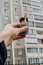 A hand holds an open matchbox with several burnt matches protruding from it. In the background, there is a multi-story residential building with large windows and a faded facade.