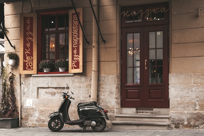 A small black scooter is parked in front of a charming cafe or restaurant with a rustic stone exterior. The windows are adorned with plants in white pots, and the door and window frames are painted in a deep maroon color. The cafe signs display the words 'cafe' and 'pizza napoletana' in elegant script on wooden panels.