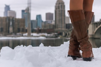 Brown leather boots stand in the snow with a cityscape in the background. Skyscrapers and buildings rise against a grey, cloudy sky while a river, partially covered with snow, separates the boots from the urban skyline.