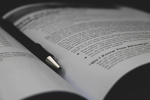 An open book with printed text visible on the pages, accompanied by a black pen resting diagonally across one of the pages. The text includes bullet points and appears to be educational material.