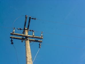A utility pole with several wires and insulators set against a clear blue sky. The structure is constructed of wood and metal components, with multiple cables extending outward.