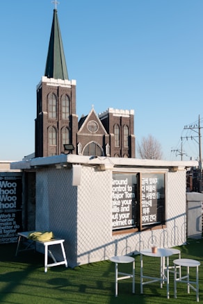 A black Gothic-style church with tall spires and arched windows stands prominently against a clear blue sky. In the foreground, there is a roof area with green artificial turf, featuring modern outdoor furniture including a small table, two chairs, and a bench with cushions. The side of the building has large windows with text in English and another language, possibly Korean.