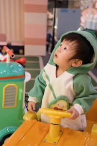 A young child is wearing a green and white costume with a hood, standing in a play area. The child is holding a yellow steering wheel, looking upward with curiosity. Nearby, there is a brightly colored play structure featuring vibrant hues of green and yellow.