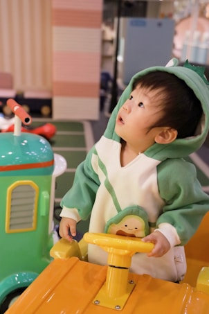 A young child is wearing a green and white costume with a hood, standing in a play area. The child is holding a yellow steering wheel, looking upward with curiosity. Nearby, there is a brightly colored play structure featuring vibrant hues of green and yellow.