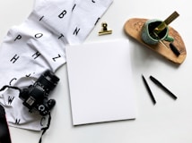 A flat lay of various items includes a camera, a white cloth with black letters, a clip, a blank white sheet, two black pens, a cup with a paintbrush and ruler, and a wooden tray. The arrangement is aesthetically pleasing with a clean and minimalistic setup.