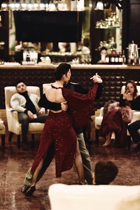 A couple is dancing in the center of a dimly lit lounge area. The woman is wearing a red dress with an open back and the man is in dark clothing. Several seated individuals are watching the performance, some sitting casually in chairs, while others appear to be more engaged. Behind them, a bar counter with various items like bottles is visible.