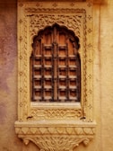 An ornate wooden window with intricate carvings set within a decorative stone frame. The wood appears aged, with dark metal accents creating a grid pattern. The stone frame features floral and geometric motifs, enhancing its antique appearance.