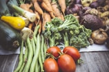 An assortment of fresh vegetables laid out on a wooden surface, including zucchini, yellow squash, green beans, broccoli, tomatoes, carrots, onions, and beets. The vegetables are arranged in a visually appealing manner, emphasizing their vibrant colors and natural textures.