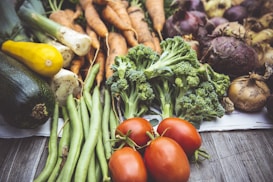 An assortment of fresh vegetables laid out on a wooden surface, including zucchini, yellow squash, green beans, broccoli, tomatoes, carrots, onions, and beets. The vegetables are arranged in a visually appealing manner, emphasizing their vibrant colors and natural textures.