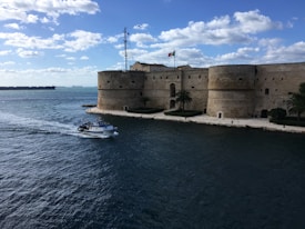 An ancient stone fortress stands beside a body of water with a small boat passing by. The building has large round towers and an Italian flag flying on top. Palm trees and well-maintained hedges are visible along the shoreline. In the background, the open sea stretches out under a partly cloudy sky.