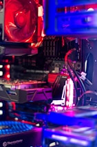 A close-up view of a computer's internal components, including a graphics card, cooling fan, and various cables. The setup features vibrant LED lighting in red, purple, and blue hues, highlighting the detailed structure of the hardware.
