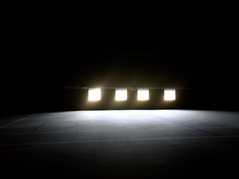 A dimly lit indoor sports court with a polished floor. Four rectangular light sources illuminate the central area, casting subtle shadows. The surroundings are mostly dark, creating a high contrast with the bright windows.
