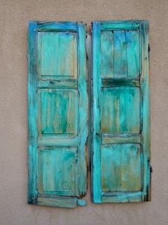 Two rustic, weathered turquoise wooden shutters are mounted on an aged beige wall. The shutters have a textured, rough surface with visible wood grain and wear, providing a vintage and rustic appearance.