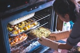 A person wearing oven mitts is carefully placing a baking dish filled with mixed vegetables into an open oven. The oven contains multiple trays of roasted food, including dishes of potatoes and other vegetables. The setting appears to be a home kitchen.