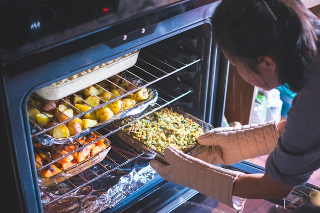 A person wearing oven mitts is carefully placing a baking dish filled with mixed vegetables into an open oven. The oven contains multiple trays of roasted food, including dishes of potatoes and other vegetables. The setting appears to be a home kitchen.