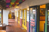 A rustic corridor with colorful papel picado banners hanging from the ceiling. The area features yellow doors and walls with a southwestern architectural style. Signs on the door advertise tamales, elotes, and champurrado. Outdoor seating is visible with wooden tables and chairs, adding to the cozy atmosphere.