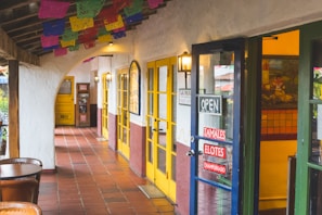 A rustic corridor with colorful papel picado banners hanging from the ceiling. The area features yellow doors and walls with a southwestern architectural style. Signs on the door advertise tamales, elotes, and champurrado. Outdoor seating is visible with wooden tables and chairs, adding to the cozy atmosphere.