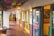 A rustic corridor with colorful papel picado banners hanging from the ceiling. The area features yellow doors and walls with a southwestern architectural style. Signs on the door advertise tamales, elotes, and champurrado. Outdoor seating is visible with wooden tables and chairs, adding to the cozy atmosphere.