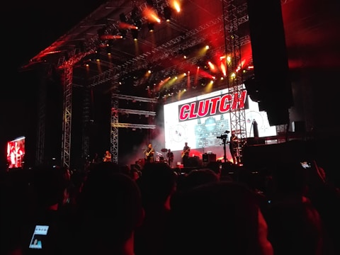 A live concert scene with a band performing on a large stage. The stage is illuminated with red and warm white lights, creating a dramatic effect. A large digital screen behind the band displays graphics and the word 'CLUTCH'. The audience stands closely packed, some capturing the performance on their phones.