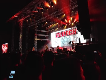 A live concert scene with a band performing on a large stage. The stage is illuminated with red and warm white lights, creating a dramatic effect. A large digital screen behind the band displays graphics and the word 'CLUTCH'. The audience stands closely packed, some capturing the performance on their phones.