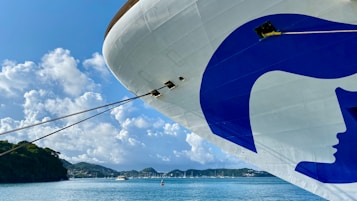 A large cruise ship with a prominent blue and white logo on its bow is docked in a serene harbor. The background features a clear blue sky with fluffy clouds and lush green hills. Several smaller boats are moored in the distance on the calm blue water.