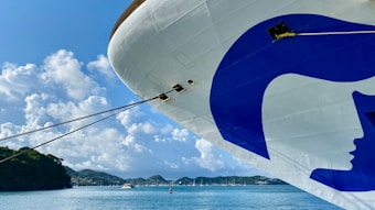 A large cruise ship with a prominent blue and white logo on its bow is docked in a serene harbor. The background features a clear blue sky with fluffy clouds and lush green hills. Several smaller boats are moored in the distance on the calm blue water.