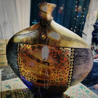 An ornate, large decorative vase with a wooden appearance and intricate floral designs in gold and red. The central panel features detailed embellishments and geometric patterns surrounded by metallic studs. It's placed on a patterned surface with a background of draped fabric and soft lights.