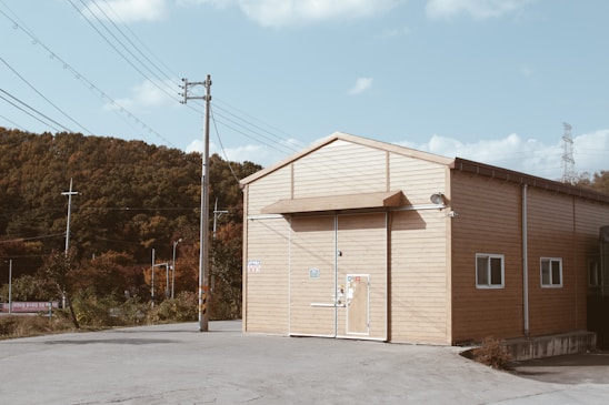 A simple, wooden, barn-like structure sits on a concrete foundation with a large door centered on the front side. The building is surrounded by a clear sky and power lines. In the background, a forested area adds an element of nature contrasting with the man-made environment.