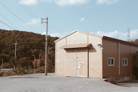 A simple, wooden, barn-like structure sits on a concrete foundation with a large door centered on the front side. The building is surrounded by a clear sky and power lines. In the background, a forested area adds an element of nature contrasting with the man-made environment.