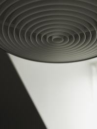The image features a close-up of a circular structure with concentric rings creating a pattern. It is likely part of an architectural or interior design element with a minimalist aesthetic. The lighting is soft, casting shadows that enhance the depth and dimension of the rings.