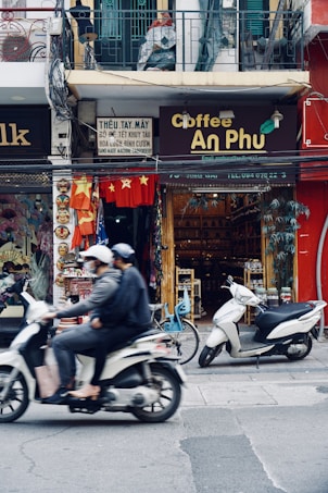 A bustling street scene capturing two individuals on a moving motorcycle in front of a small coffee shop. The coffee shop, named 'Coffee An Phu', is situated between other shops with a variety of items on display, including souvenirs and colorful decorations. A second motorcycle and a bicycle are parked nearby. The upper level shows a balcony with a mannequin and some textiles hanging.
