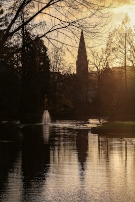 A serene landscape with a lake reflecting the warm glow of a sunset. Bare trees frame a church steeple in the background. The water is calm, with a small fountain creating gentle ripples.
