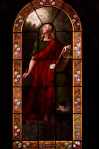 A stained glass window illustrates a figure draped in a red and green robe, holding a scroll. The figure appears poised and contemplative, with long, curly blond hair. The background features cloud-like designs, and the border is adorned with intricate floral patterns in vibrant colors.