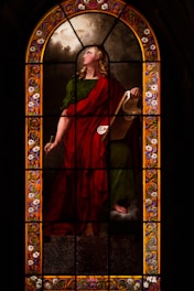 A stained glass window illustrates a figure draped in a red and green robe, holding a scroll. The figure appears poised and contemplative, with long, curly blond hair. The background features cloud-like designs, and the border is adorned with intricate floral patterns in vibrant colors.