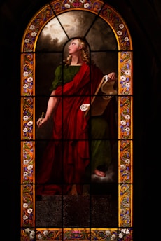 A stained glass window illustrates a figure draped in a red and green robe, holding a scroll. The figure appears poised and contemplative, with long, curly blond hair. The background features cloud-like designs, and the border is adorned with intricate floral patterns in vibrant colors.