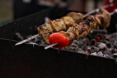 Two skewers of meat and vegetables, with a focus on a red tomato, are being grilled over hot charcoal. The coals are glowing with heat, and the skewers appear to be cooking slowly, with some grill marks visible on the meat.
