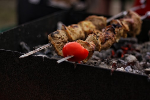 Two skewers of meat and vegetables, with a focus on a red tomato, are being grilled over hot charcoal. The coals are glowing with heat, and the skewers appear to be cooking slowly, with some grill marks visible on the meat.
