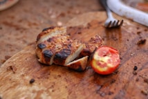 Grilled chicken pieces lie on a wooden cutting board alongside a sliced, charred tomato. The surface is slightly worn and contains scattered food crumbs, with a metal fork partially visible in the background.