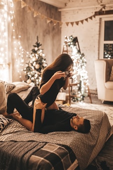 A cozy and warmly lit room features a couple engaged in a playful pose on a bed, surrounded by festive Christmas decorations. String lights adorn the walls and a decorated Christmas tree is visible in the background, adding to the intimate atmosphere. A woman is holding a camera, capturing a moment with her partner, who is lying on the bed.