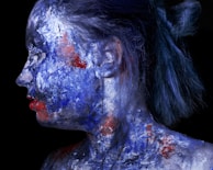 A person in profile view with the skin covered in an abstract mix of blue, white, and red paint, creating a striking contrast against a dark background. The hair is styled in a bun, and the facial features are partially obscured by the paint.