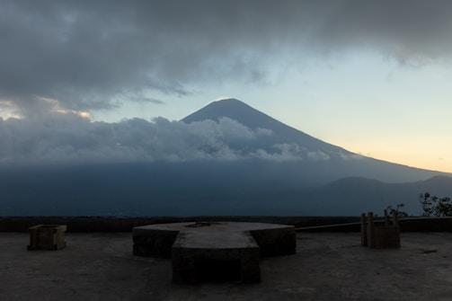A large mountain rising above a layer of clouds, with the sky transitioning from light blue to a subtle yellow near the horizon. The foreground includes a concrete platform with several angular shapes and a few small plants.