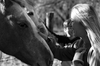 A black and white photo featuring a woman smiling as she gently touches the face of a horse. There is another person in the background, and they are all outdoors in a wooded area.