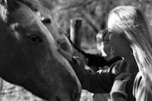 A black and white photo featuring a woman smiling as she gently touches the face of a horse. There is another person in the background, and they are all outdoors in a wooded area.