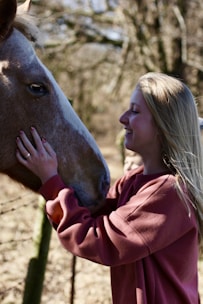 A person with long blonde hair is smiling while gently touching the face of a brown horse. They appear to be in a natural outdoor setting with blurred trees in the background.