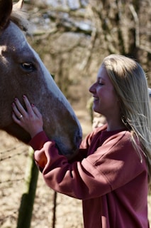 A person with long blonde hair is smiling while gently touching the face of a brown horse. They appear to be in a natural outdoor setting with blurred trees in the background.
