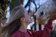 A woman with long hair is gently facing a horse, showing a moment of affectionate interaction. The horse has a spotted muzzle, and the woman wears a red sweater. They are outdoors, with trees and a blurred background.