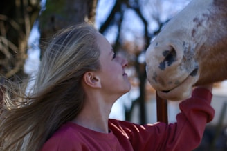 A woman with long hair is gently facing a horse, showing a moment of affectionate interaction. The horse has a spotted muzzle, and the woman wears a red sweater. They are outdoors, with trees and a blurred background.