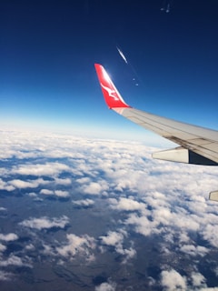 An airplane wing with a red tip and logo is visible against a backdrop of a deep blue sky. Below, a layer of fluffy white clouds stretches across the horizon, partially covering the landscape below.
