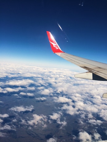 An airplane wing with a red tip and logo is visible against a backdrop of a deep blue sky. Below, a layer of fluffy white clouds stretches across the horizon, partially covering the landscape below.
